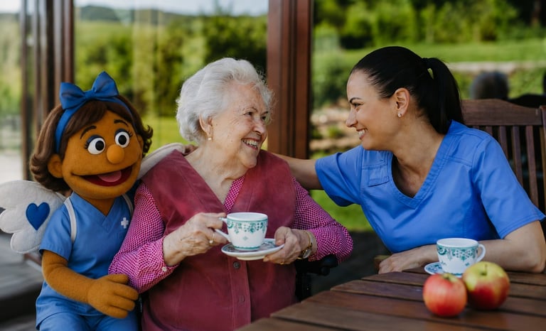 An elderly woman and a caregiver in blue scrubs smile while sharing tea with a puppet character, seated at a table near a garden window.