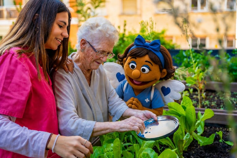 An older woman and younger woman holding a blue puppet character while examining a potted plant together in an outdoor garden setting