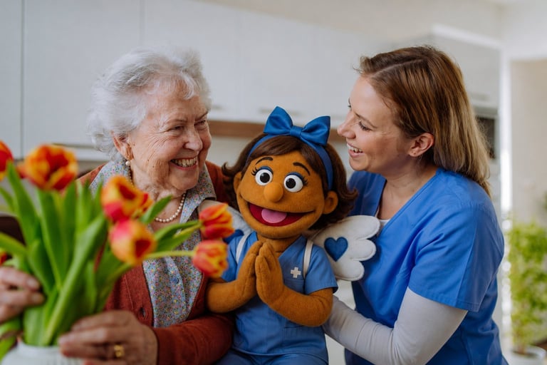 An elderly woman and a caregiver in blue hold a brown puppet with a blue bow between them, with colorful flowers in the background.