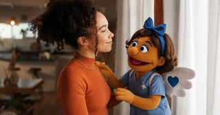 Woman in orange shirt smiling while interacting with a blue puppet character wearing a bow in a home setting